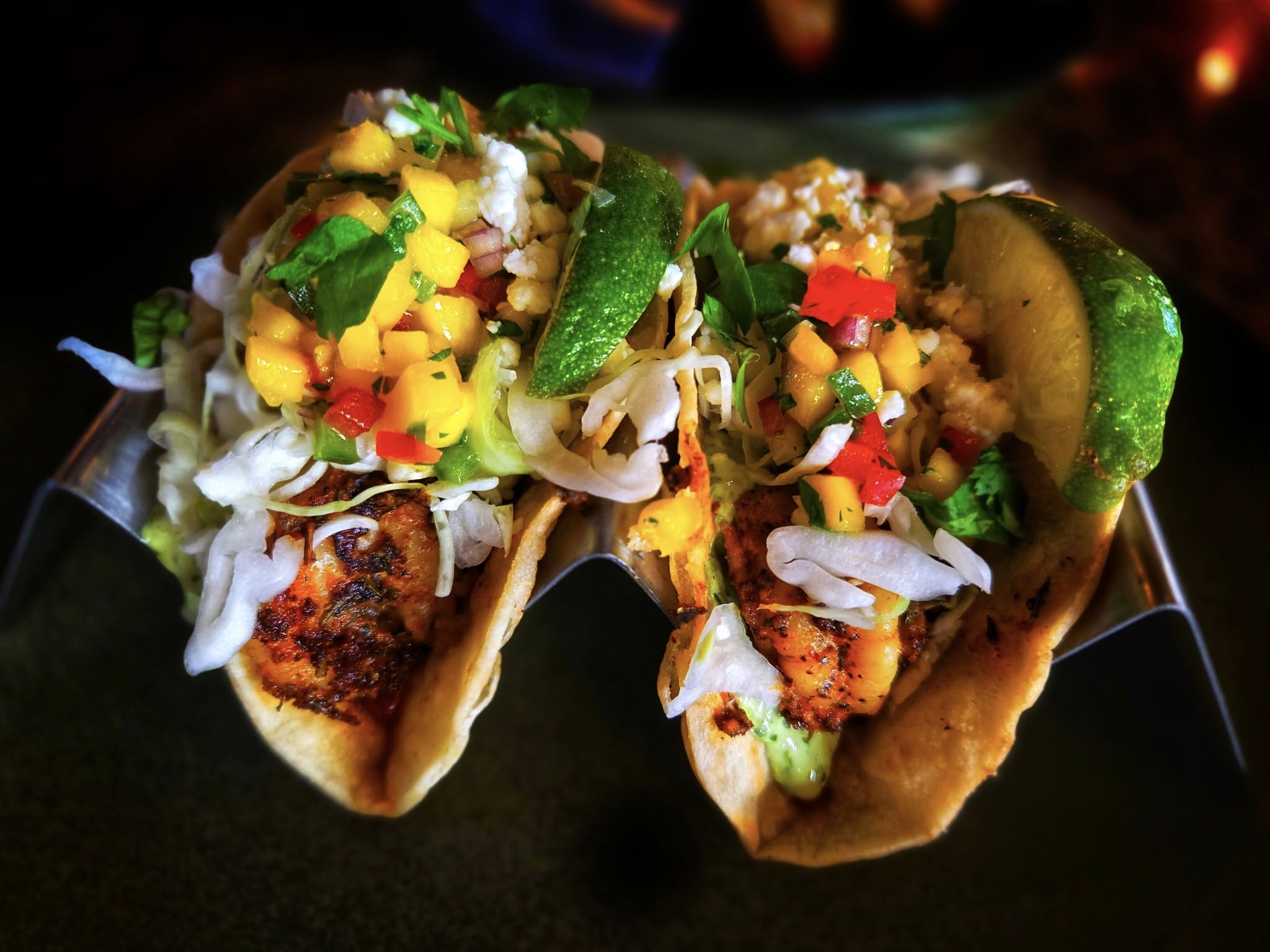 Two grilled fish tacos in soft tortillas, topped with shredded lettuce, mango salsa, chopped cilantro, and lime wedges—served in a metal taco holder. A fresh and flavorful choice from our menu.