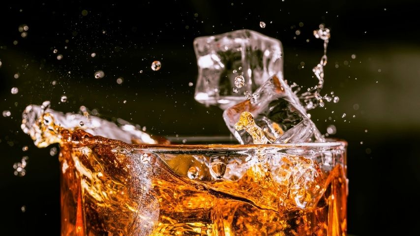 A close-up of a glass filled with amber liquid from Peach Street Distillers, as ice cubes splash into it, sending droplets flying against a dark background—a vivid capture celebrating craft spirits.