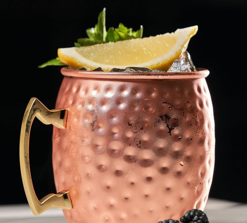 A copper mug filled with a cocktail, garnished with a lemon wedge, mint, and blackberries, sits on a white surface. Text reads New Year New Cocktails and Peach Street Distillers—featuring New Cocktails for January.