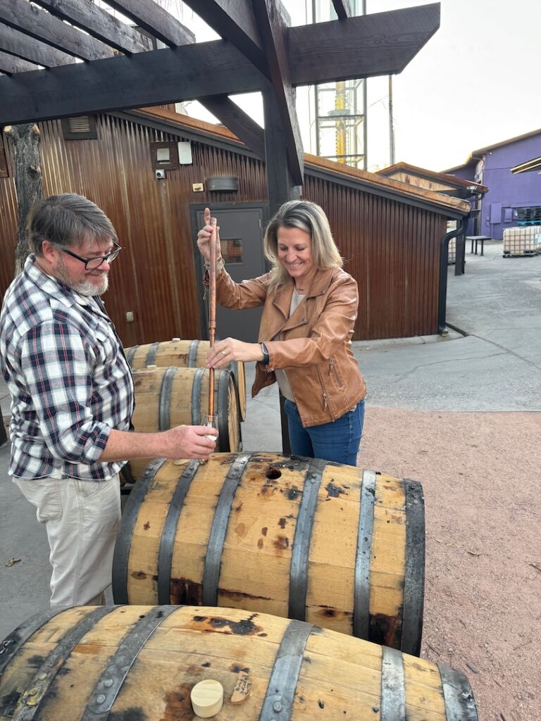 A woman uses a wine thief to extract liquid from a wooden barrel at Peach Street Distillers while a man watches, both standing outdoors near metal-clad buildings and additional barrels at the craft spirits distillery.