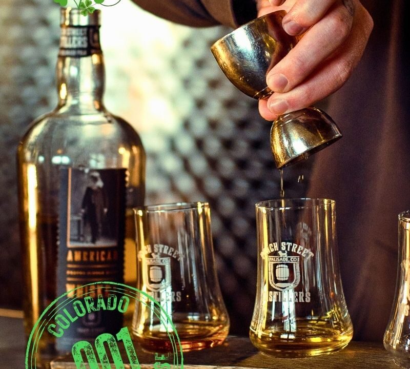 A hand pours whiskey from a jigger into a glass beside a Peach Street Distillers bottle. Shamrocks and SLÁINTE! are overlaid, celebrating Irish Spirit, with a green Colorado 001 Distiller License stamp.