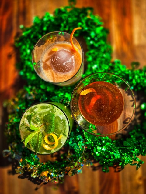 Three cocktails with citrus peels and mint, crafted with Irish Spirit, are arranged on a wooden table and surrounded by sparkling green decorations, suggesting a festive St. Patrick’s Day theme.