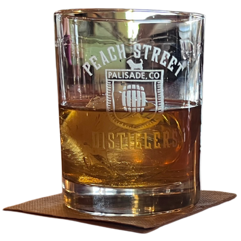 A glass of whiskey with ice sits on a brown napkin. The glass is etched with the words Peach Street Distillers, Palisade, CO and a logo featuring a peach and barrel—true craftsmanship from this Colorado spirits craft distillery.