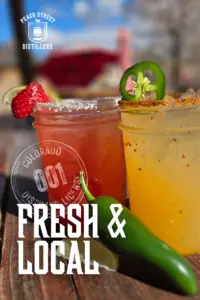 Two colorful cocktails in mason jars, one red with a strawberry garnish and one yellow with a jalapeño garnish, sit on a sunny outdoor table; text reads “Fresh & Local - Peach Street Distillers.