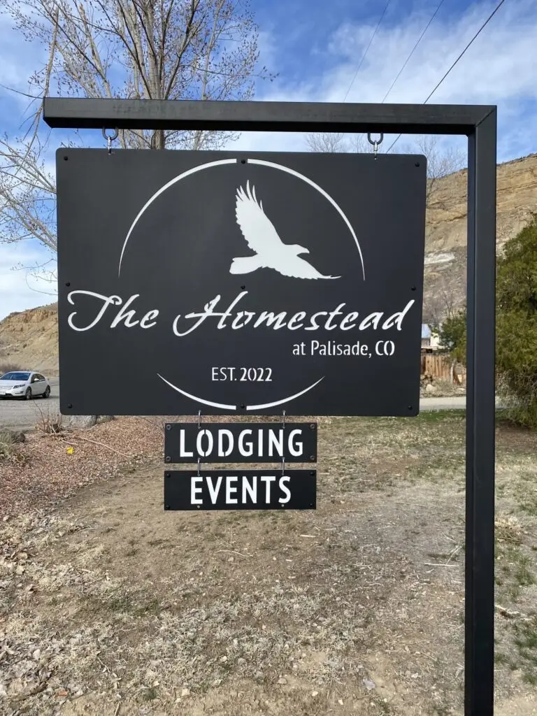 A black sign reads The Homestead at Palisade, CO with a flying bird graphic above. Smaller text says Est. 2022. Below, two separate signs read Lodging and Events—an inviting spot for your next Sip & Stay travel deal. Outdoor background with trees and hills.