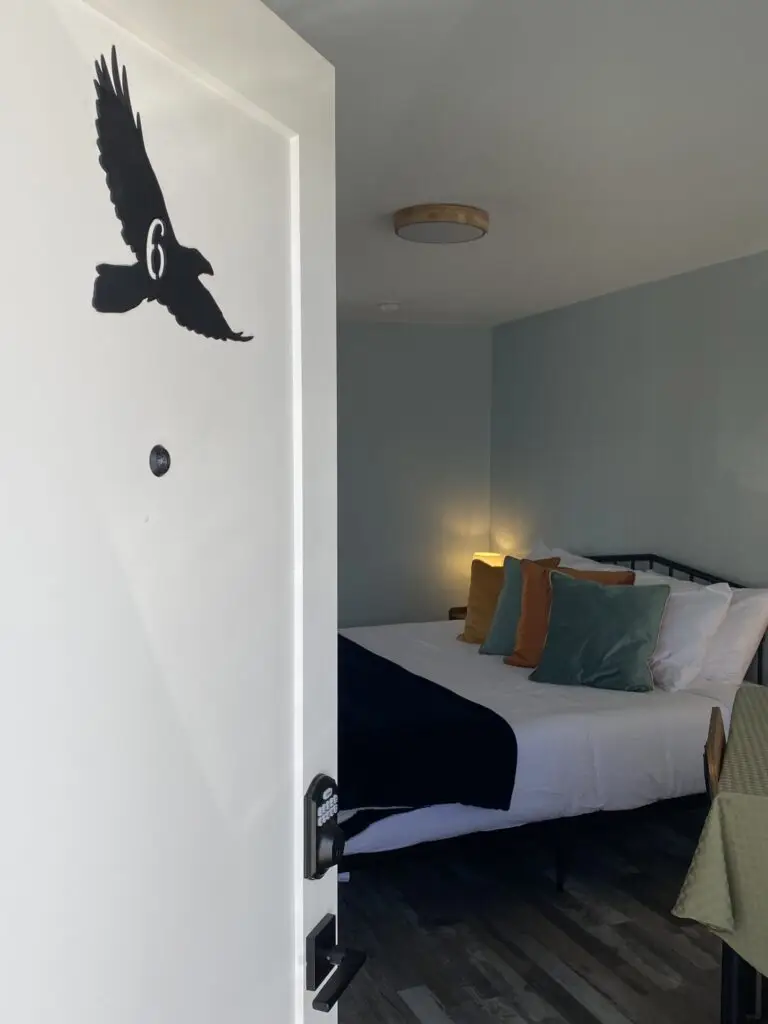 A white door with a black silhouette of a bird and the number 6 opens into a cozy bedroom—perfect for your hotel special—featuring a made bed, colorful pillows, a dark blanket, and soft lighting.
