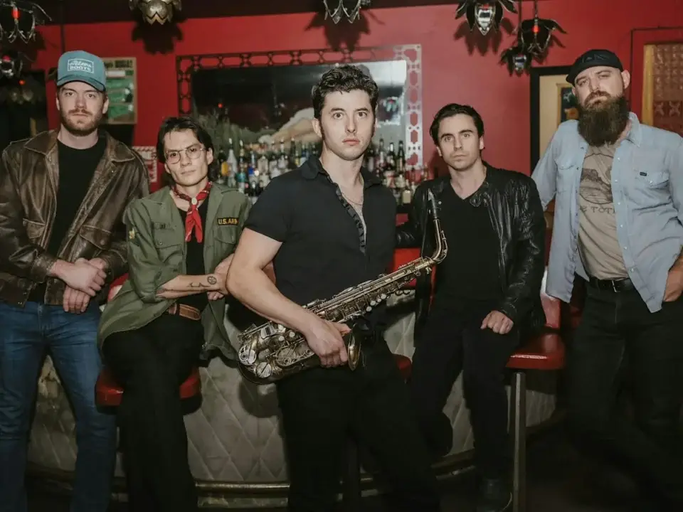 Five people pose in front of a bar with a red wall and mirror. The person in the center holds a saxophone. Dressed casually, the group creates a relaxed, vintage vibe—perfect for enjoying live music at Peach Street Distillers.