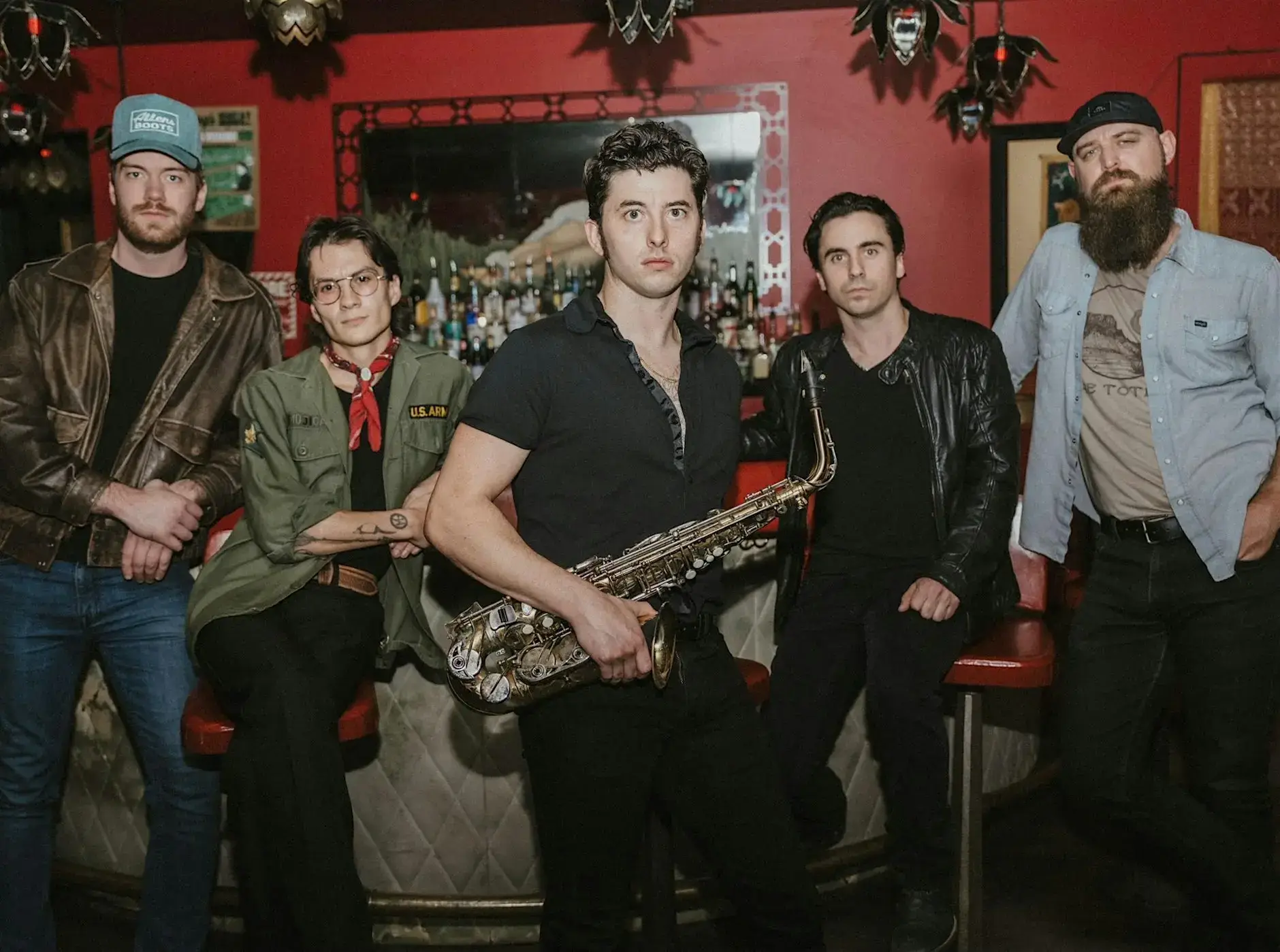Five people pose in front of a bar with a red wall and mirror. The person in the center holds a saxophone. Dressed casually, the group creates a relaxed, vintage vibe—perfect for enjoying live music at Peach Street Distillers.