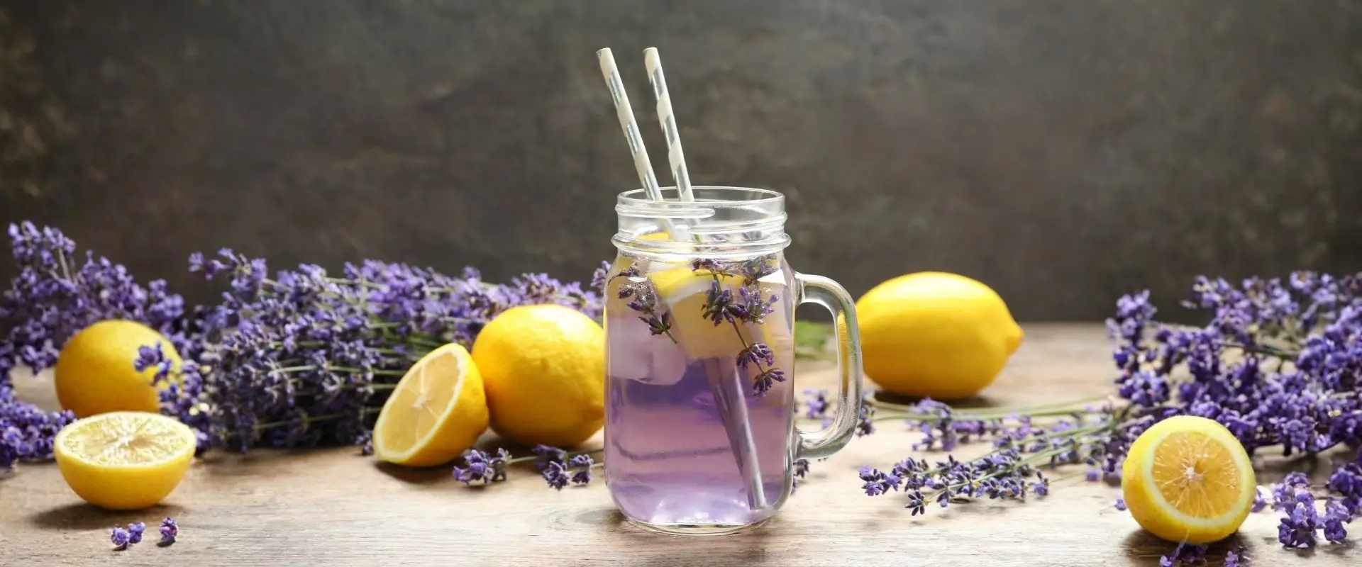 A mason jar with purple lavender lemonade, ice, and lemon slices sits on a wooden surface, surrounded by fresh lemons and sprigs of lavender—a perfect refreshment inspired by Lavender Festival drink specials.