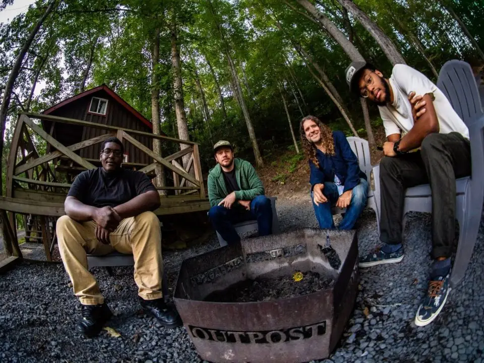 Four men sit around a fire pit labeled OUTPOST in front of a wooden cabin, surrounded by trees. The group appears relaxed, enjoying a moment outdoors on gravel near the forest as soft live music by John Inghram plays in the background.