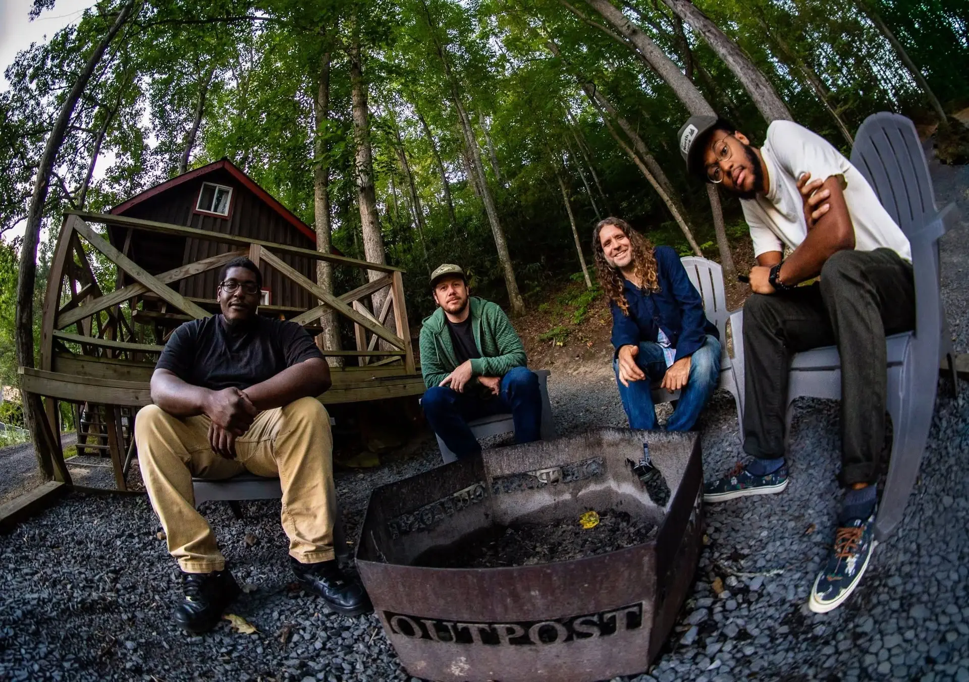 Four men sit around a fire pit labeled OUTPOST in front of a wooden cabin, surrounded by trees. The group appears relaxed, enjoying a moment outdoors on gravel near the forest as soft live music by John Inghram plays in the background.