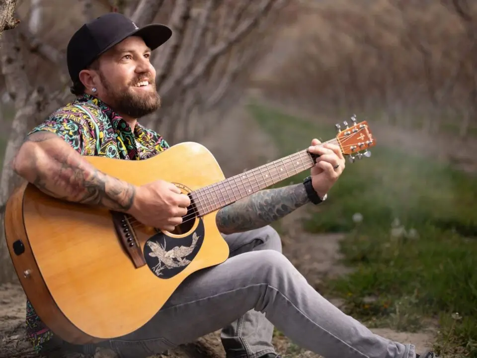 A smiling man with tattoos, wearing a colorful shirt and black cap, sits on the ground outdoors, playing acoustic guitar among rows of trees on a cloudy day—Geoff Crumbaugh shares free live music in this serene setting.