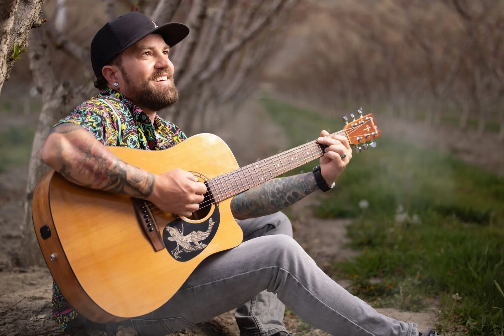 A smiling man with tattoos, wearing a colorful shirt and black cap, sits on the ground outdoors, playing acoustic guitar among rows of trees on a cloudy day—Geoff Crumbaugh shares free live music in this serene setting.