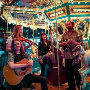 Four musicians with string instruments pose on a bright, colorful carousel with lights and painted horses. The group, known as Mama Said String Band, includes a guitarist, violinist, upright bassist, and mandolin player—all smiling at the camera and ready to share the joy of live music.