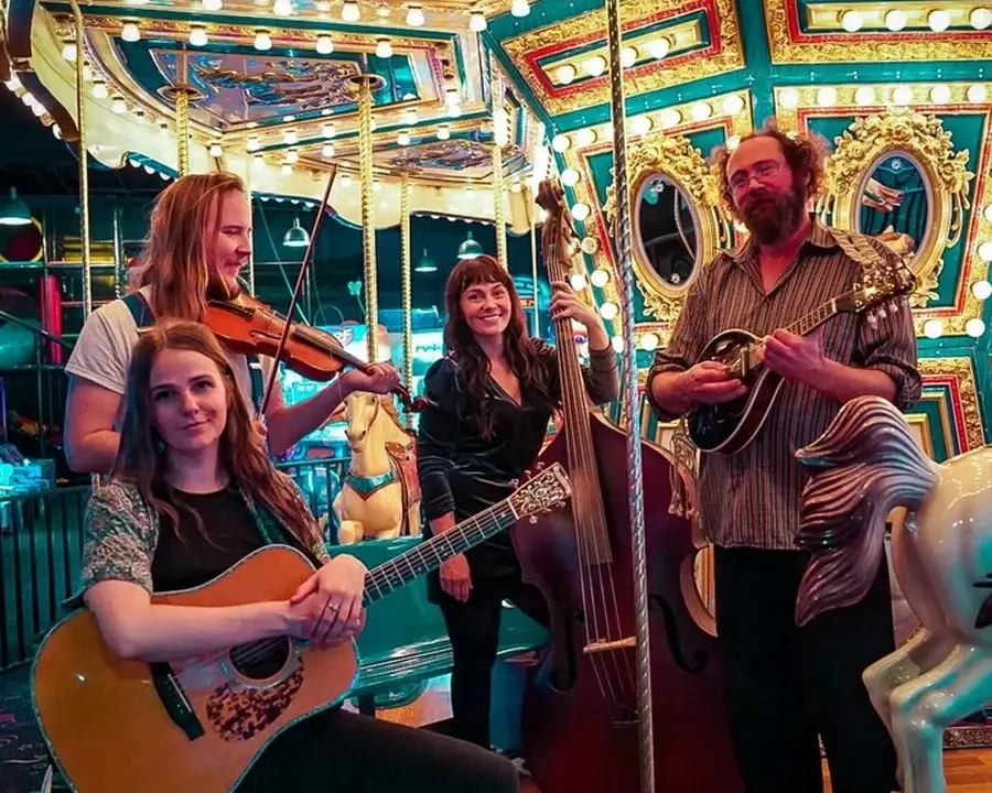 Four musicians with string instruments pose on a bright, colorful carousel with lights and painted horses. The group, known as Mama Said String Band, includes a guitarist, violinist, upright bassist, and mandolin player—all smiling at the camera and ready to share the joy of live music.