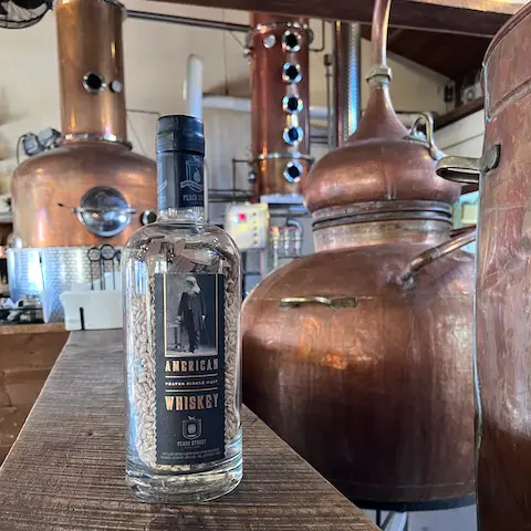 A bottle of American whiskey stands on a wooden surface in front of large copper distillery equipment inside a distillery, inviting visitors to explore the rich flavors this spirit brings to any tasting menu.