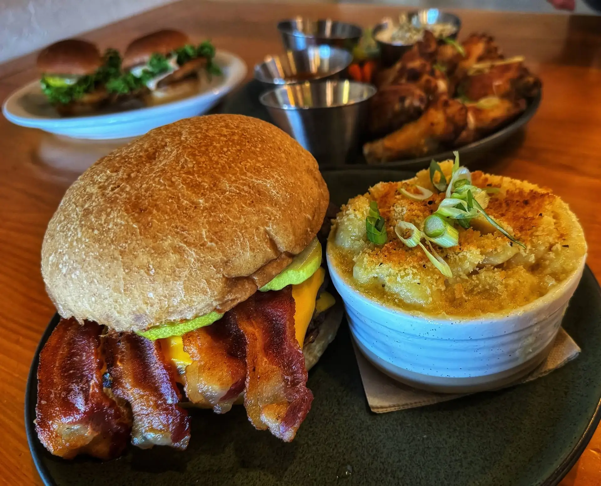 A plate with a bacon cheeseburger and a bowl of baked macaroni and cheese sits on a wooden table, inviting you to savor classic comfort food. In the background, sliders and other dishes are slightly out of focus—just a glimpse of the delicious offerings you’ll find when you visit Peach Street Distillers’ welcoming distillery.