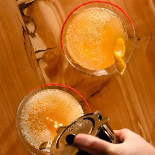 Two orange cocktails on a wooden table, one garnished with a citrus twist. A hand holds a cocktail shaker, pouring a drink into one of the glasses. The scene is warmly lit from above.