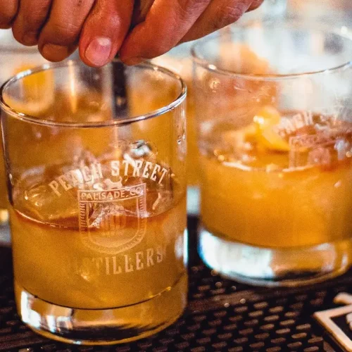 Two glasses of an orange-colored cocktail with ice, one being stirred by a hand. Both glasses have Peach Street Distillers printed on them and sit on a bar mat, highlighting a refreshing choice from the menu.